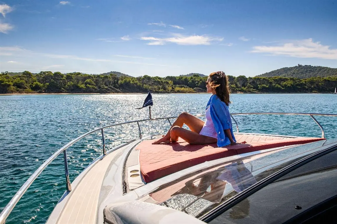 Woman lounging on a luxury boat's bow, enjoying sunshine and water views, with lush greenery in the background.
